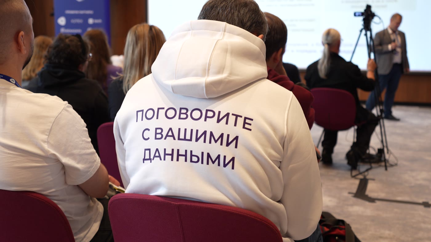 Репортажная съёмка форума «Управление данными — 2026»: участник в худи «Поговорите с вашими данными» в зале, на заднем плане — камера на штативе